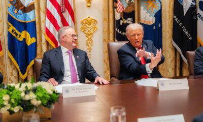 Prime Minister Anthony Albanese and President Donald J. Trump seated at a meeting table in the White House, engaged in conversation during their first official bilateral meeting, with military flags in the background.
