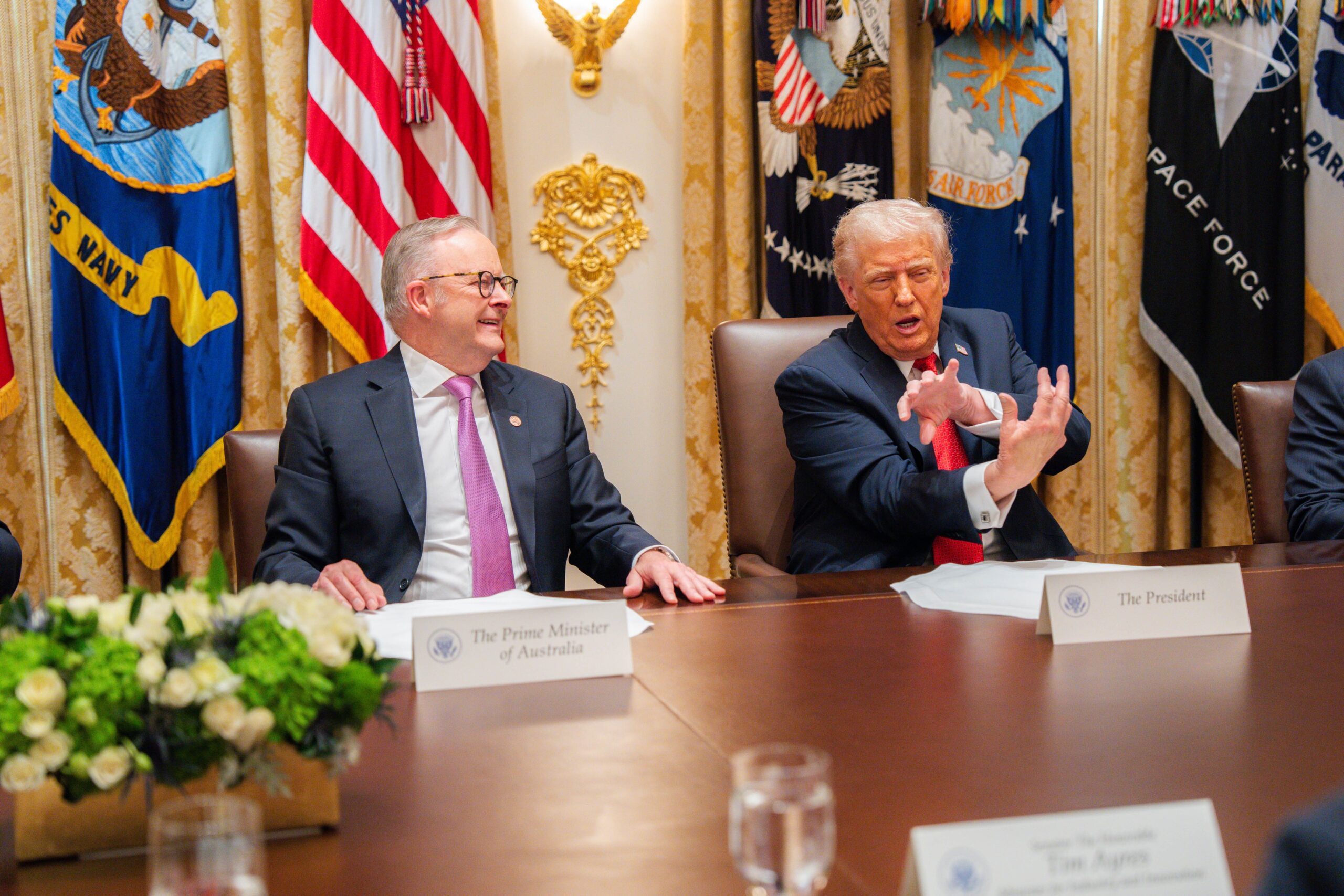Prime Minister Anthony Albanese and President Donald J. Trump seated at a meeting table in the White House, engaged in conversation during their first official bilateral meeting, with military flags in the background.