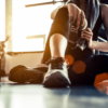Sport woman sitting and resting after workout or exercise in fitness gym with protein shake or drinking water on floor. Relax concept. Strength training and Body build up theme. Warm and cool tone