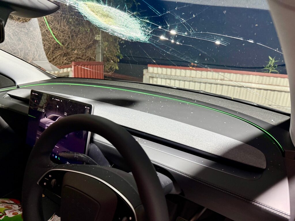 Interior view of a Tesla showing the damaged windshield with a melted center and crater-like impact point, glass shards visible on the dashboard.
