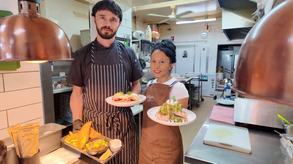 Chefs Andrew and Ayu standing in the Boston Bay Diner kitchen, holding plated dishes and a tray of fish and chips.